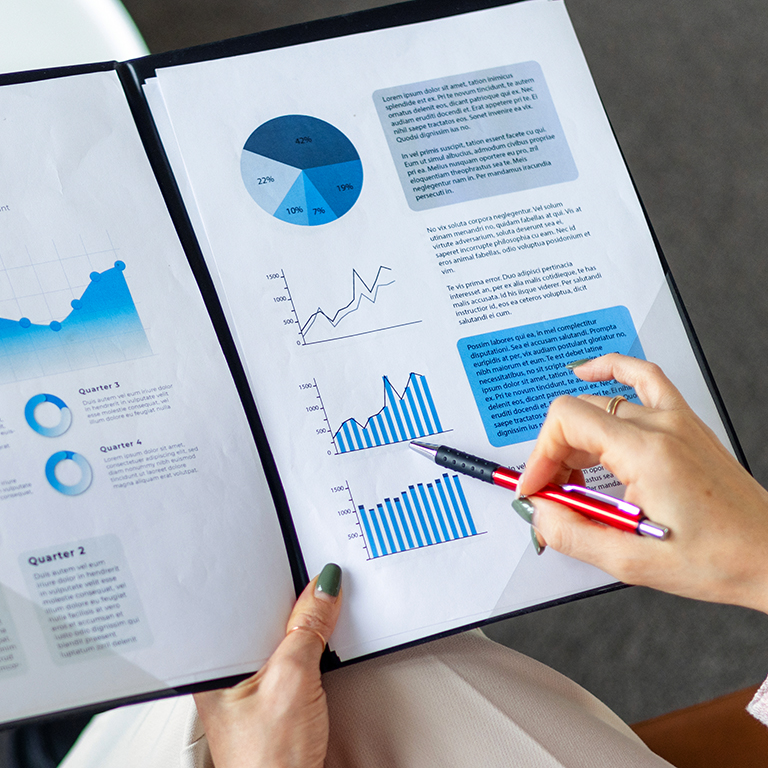 Hand holding a pen pointing at a bar graph on a document with various charts and graphs.