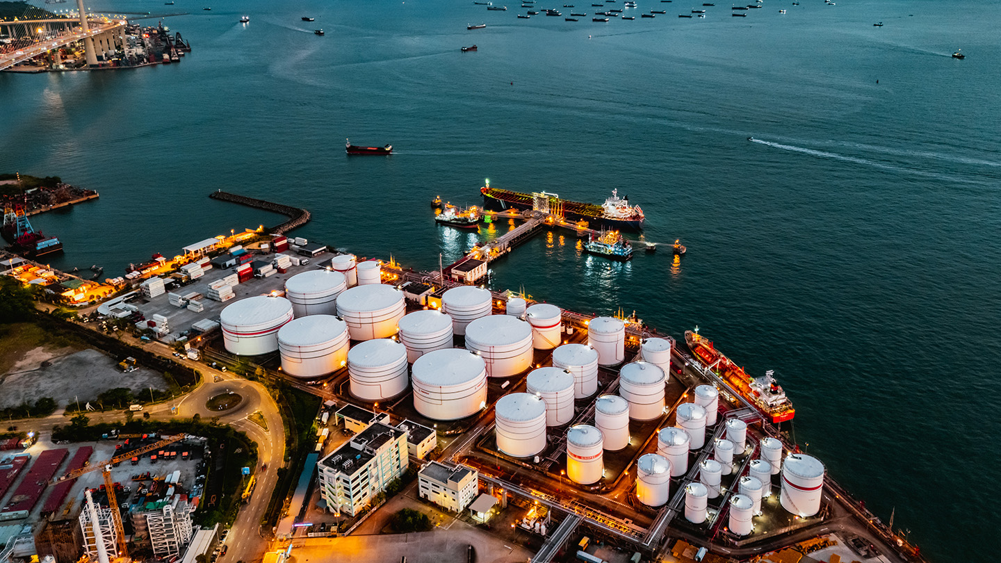Aerial view of an industrial port with large white storage tanks, docked ships, and illuminated facilities near the water. Numerous vessels are visible in the surrounding sea.